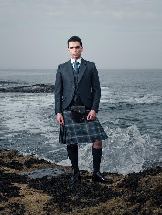 A man is standing on a rocky shore wearing Hebridean Hoolie tartan kilt outfit with waves crashing behind him
