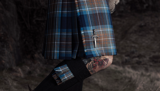 A man wearing a Holyrood Tartan Kilt and tweed jacket is standing on the slopes of Arthurs Seat in Edinburgh overlooking Holyrood Park