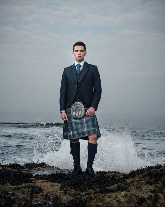 A man is standing on a rocky shore wearing Hebridean Hoolie tartan kilt outfit with waves crashing behind him