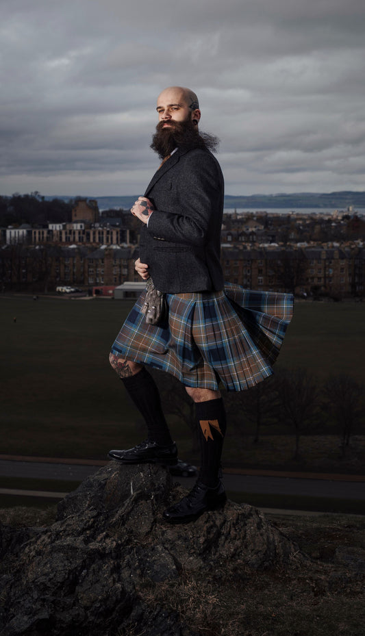 A man wearing a Holyrood Tartan Kilt and tweed jacket is standing on the slopes of Arthurs Seat in Edinburgh overlooking Holyrood Park