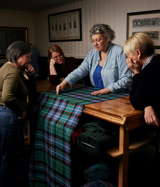 A woman is teaching aspects of tyartan cloth to three female kiltmaking students, standing round a table