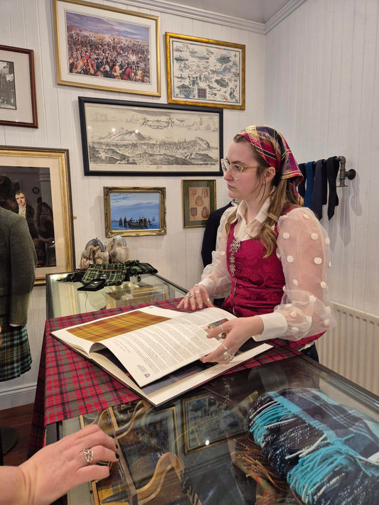 A young woman dressed in traditional Norwegian Bunad displays details of Scottish Tartan patterns and cloth examples to an unseen audinece. Historic Scottish  Illustrations are displayed on the wall behind her.