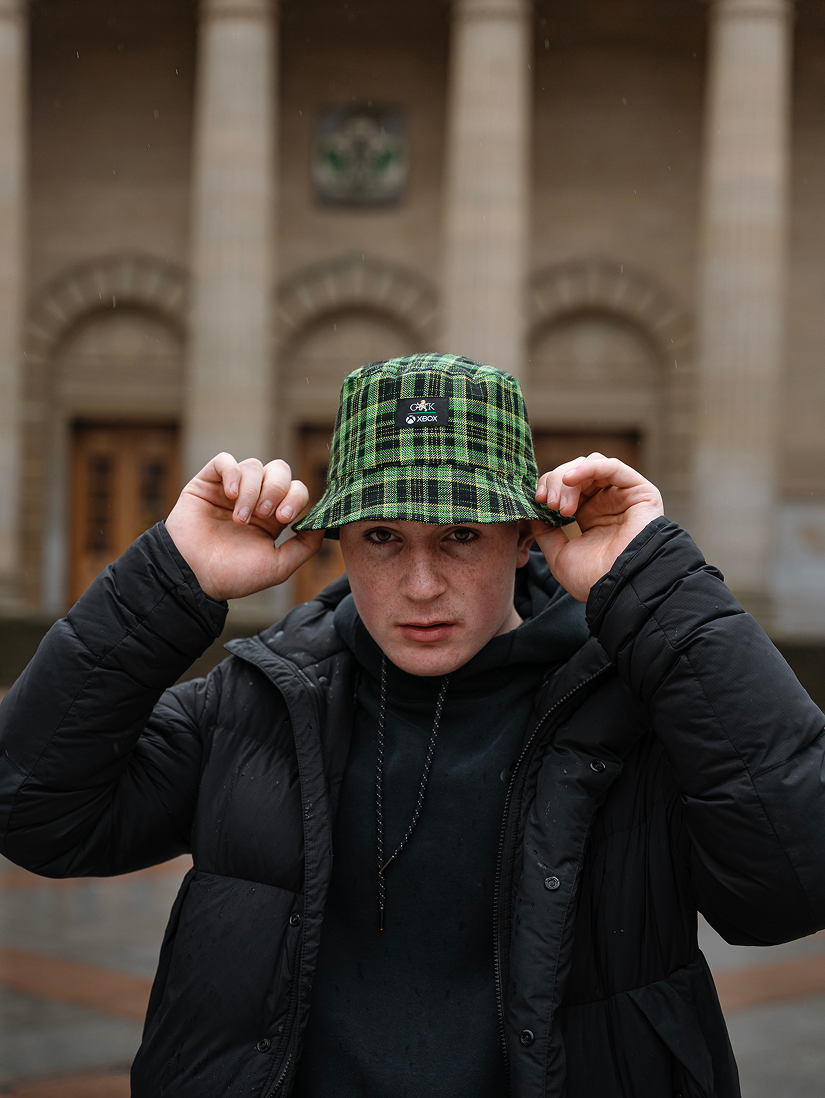 Person wearing a green plaid bucket hat and black jacket in front of a building.
